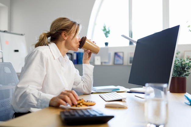 Сосредоточена на работе за компьютером, деловая женщина пьет кофе с печеньем в офисе
