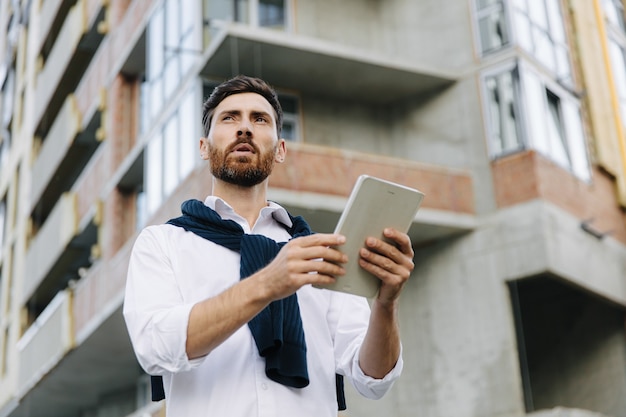 Focused young architect using digital tablet while standing at construction site. Bearded specialist with modern tablet controlling working process.