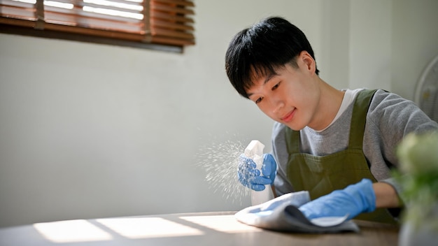 Focused young Asian man wearing rubber gloves using cleaning spray and dust cloth