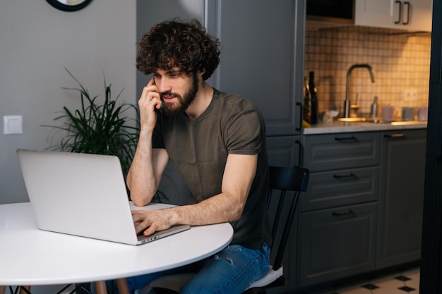 Focused young bearded business man talking on smartphone sitting at table with laptop in kitchen with modern interior looking on screen