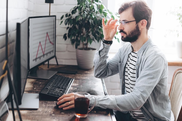 Focused young businessman in eyewear checks graphics on computer monitors Concentrated millennial man in glasses works at home on selfisolation