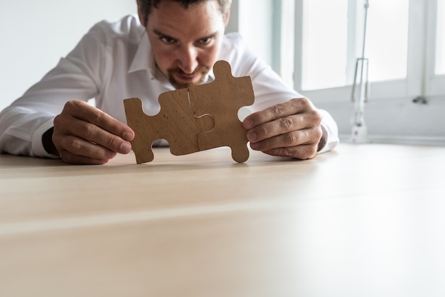 Giovane uomo d'affari concentrato che unisce due pezzi di puzzle corrispondenti mentre si siede alla sua scrivania. concettuale di fusione aziendale e soluzione.