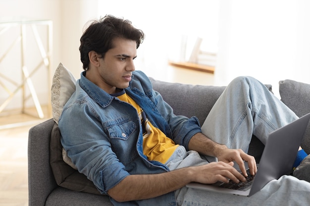 Focused young caucasian freelancer businessman lying on the couch at home with laptop working or studying remotely online