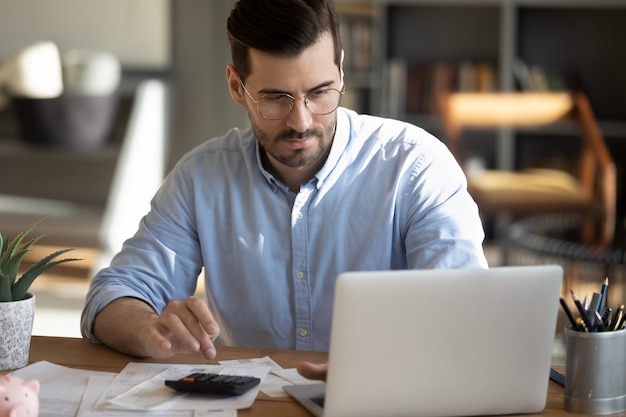 Focused young Caucasian man look at laptop screen calculate expenses expenditures pay bills taxes online Millennial male busy managing household family budget take care of financial paperwork