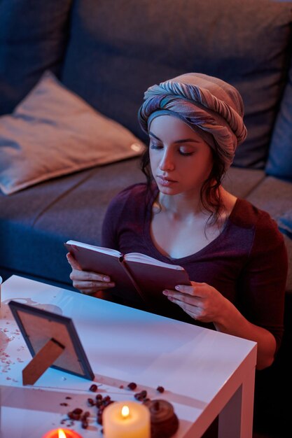 Concentrata giovane donna caucasica nel copricapo che consulta un libro prima di eseguire un rituale magico