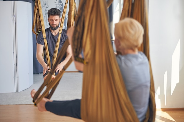 Focused young dark-haired trainer and a blonde woman sitting in silk hammocks during the workout