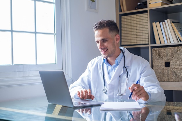 Focused young doctor wearing glasses working on laptop in office sitting at desk looking at screen serious therapist gp writing report or email consulting patient online filling medical cardxA