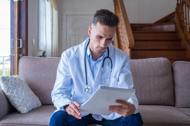 Focused young male doctor in the sofa and medical coat working with paper registry book making notes managing patients visits recording healthcare treatment sitting on couch at homexA