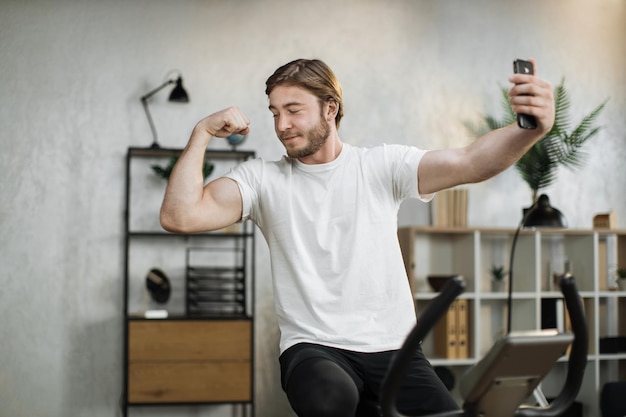 Focused young male wearing sportswear doing selfie showing biceps while using exercise bike