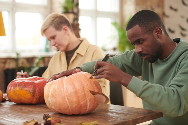 Focused young multiethnic people sitting at table and participating in pumpkin carving workshop befo