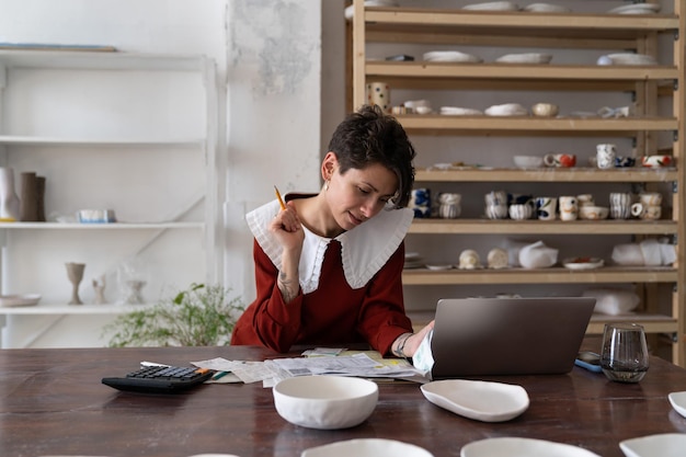 Focused young woman ceramic studio owner analyzing bills and receipts doing pottery cost analysis