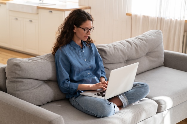 Focused young woman type message on laptop concentrated surf web sitting on couch in living room