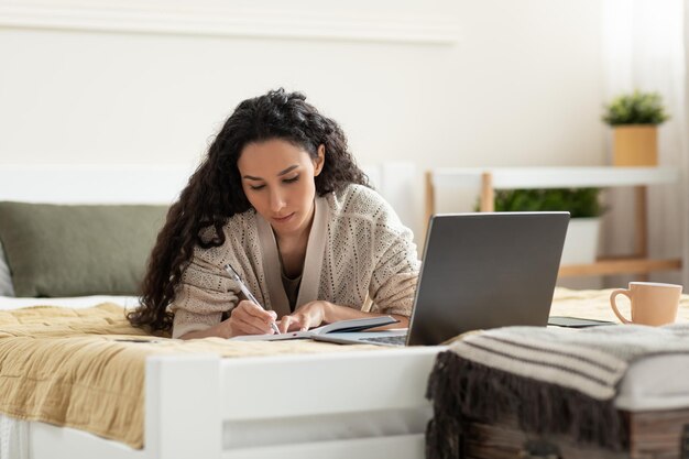 Focused young woman using laptop working or studying online taking notes during business meeting or