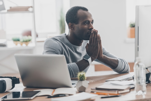 Focusing on work. Handsome young African man holding hands together while sitting at his working place in modern office