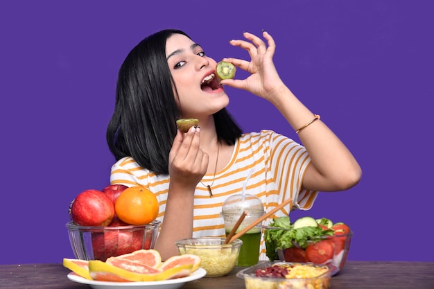 foddie girl sitting at fruit table posing over purple background eating kiwi indian pakistani model