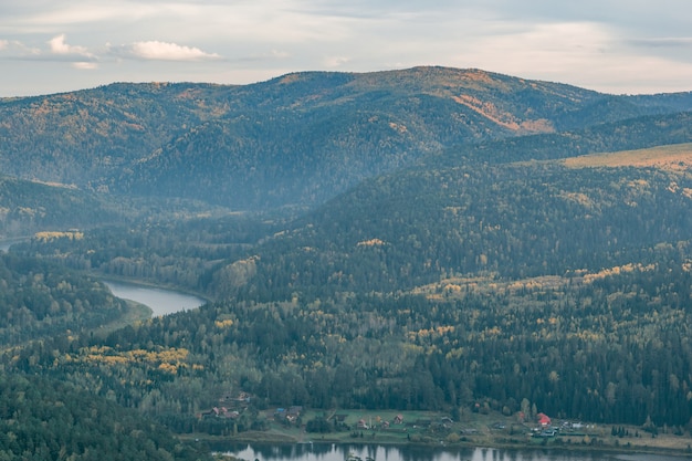 Foggy autumn landscape of the river valley on the background of mountains covered with green trees and yellow birches, natural autumn background.Bird`s-eye view.