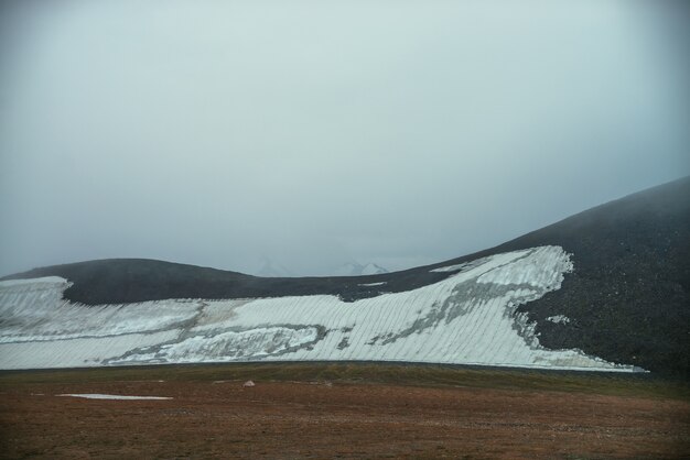 Туманный минималистский пейзаж со снежной вершиной внутри низких облаков над небольшим ледником на склоне скалистого холма в тумане. Горный туманный минимализм со снежной вершиной в облачном небе над палаткой в высокогорной долине.