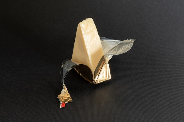 Обернутый фольгой кусочек плавленого сливочного сыра на черном фоне. Небольшой треугольный кусочек порционного мягкого сыра в золотой алюминиевой фольге.