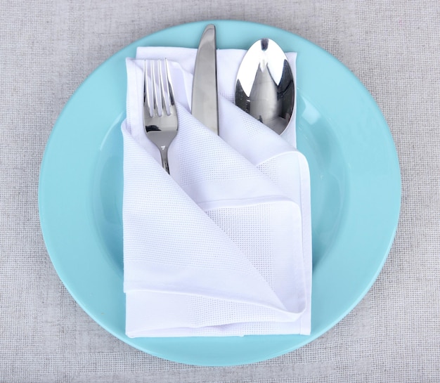 Folded napkin with fork spoon and knife on plate on color background