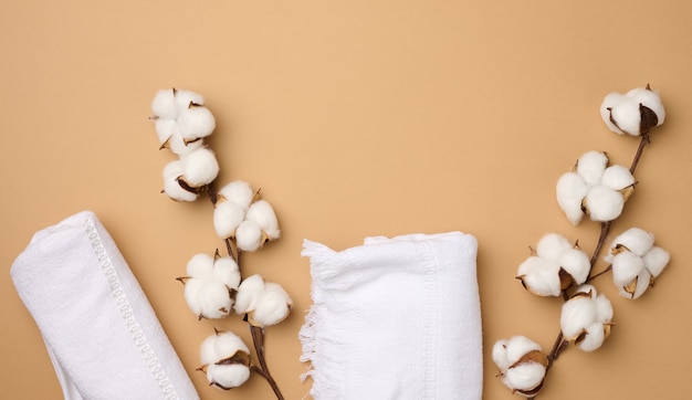 Photo folded white cotton terry towel and sprigs of cotton flower on a light brown background, top view