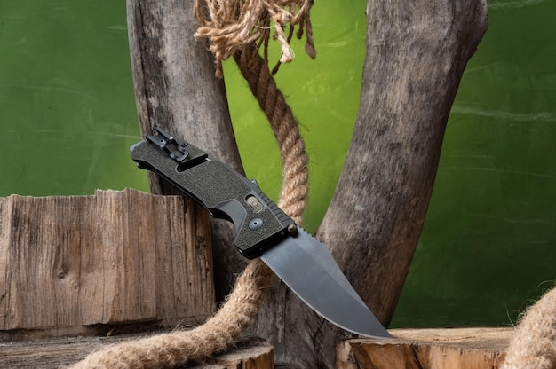 写真 黒い刃の折りたたみアーミー ナイフ 木とロープの背景にナイフ 斜めに垂直にナイフ 正面図