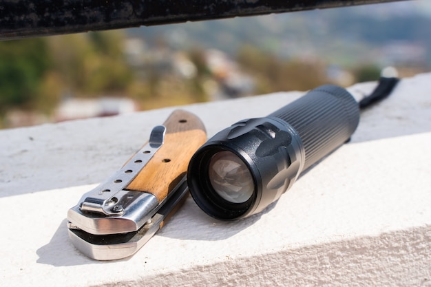 Folding knife and flashlight lying on the concrete fence. Tourist equipment, trekking, hiking and camping activity concept. Stock photo.