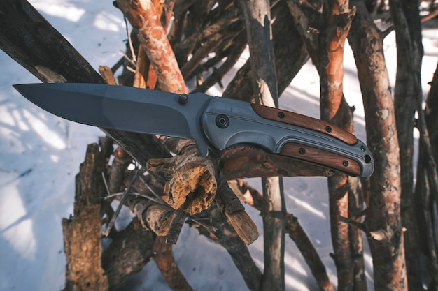 생존과 하이킹을 위한 접는 전술 칼은 소나무 겨울 숲에서 쓰러진 나무 줄기에 꽂혀 있다