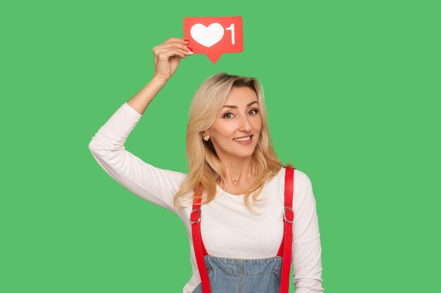 Follow and like social media blog. Portrait of lovely smiling adult woman in stylish denim overalls holding network heart icon over head, emoji notification button. indoor studio shot, isolated