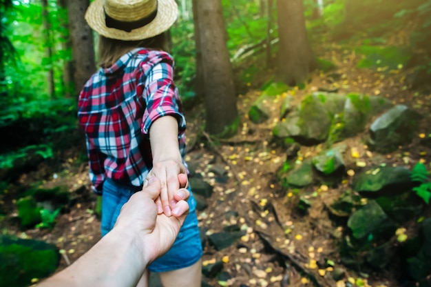 Follow me photo in the mountains. Stylish woman in checkered shirt and straw hat. Wanderlust concept. Couple hiking and travelling in summer. Holding hands in nature.