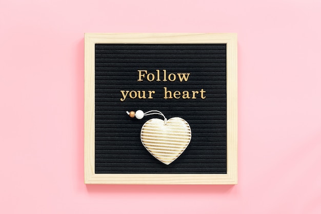 Follow your heart. Motivational quote in gold letters and decorative textile heart on black letter board on pink background.