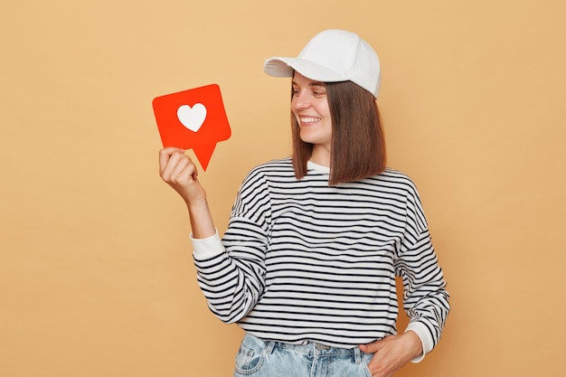 Photo following bloggers for content clicking the like button on posts smiling woman wearing striped shirt and baseball cap holding like blogger icon standing isolated over beige background