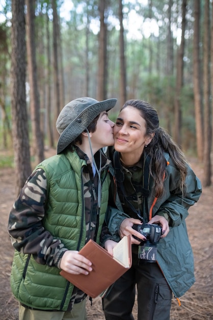 Fond mother with son with camera in forest. Schoolboy kissing mother on cheek. Hobby, photography concept