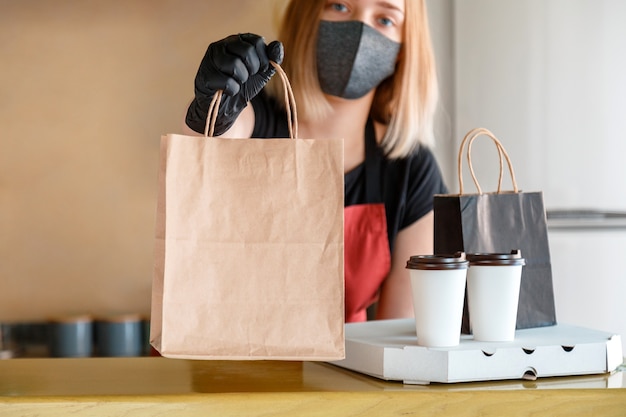 Food bag, pizza, drink package to go in takeaway restaurant. Kitchen worker issues online orders in gloves and mask. Takeaway food paper bag. Contactless food delivery lockdown covid 19.