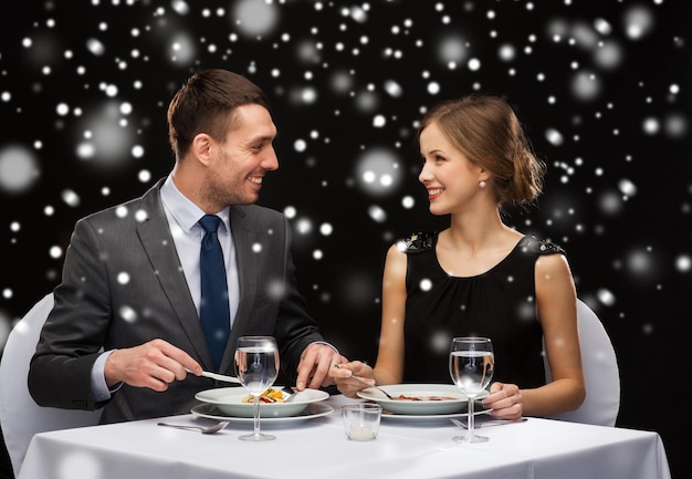 Concetto di cibo, natale, vacanze e persone - coppia sorridente che mangia portata principale al ristorante su sfondo nero innevato
