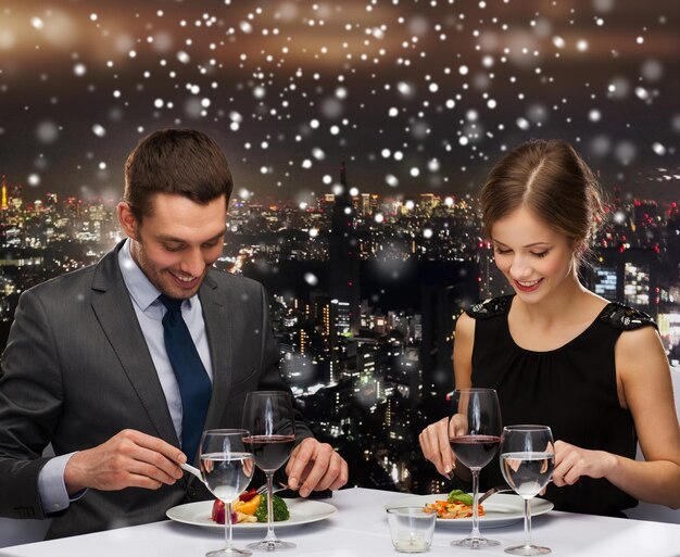 food, christmas, holidays and people concept - smiling couple eating main course at restaurant over snowy night city background