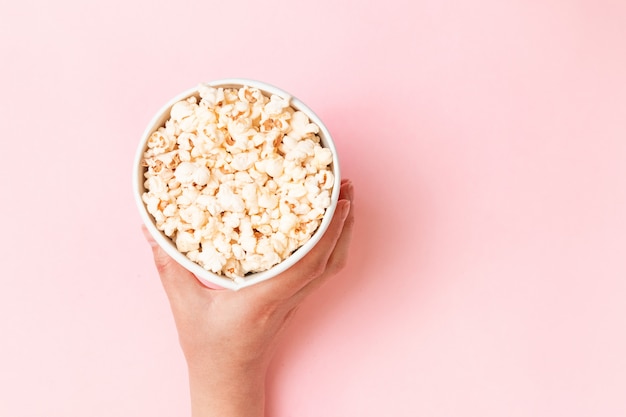 Food composition of hand holding popcorn in bucket on pink background top view copy space