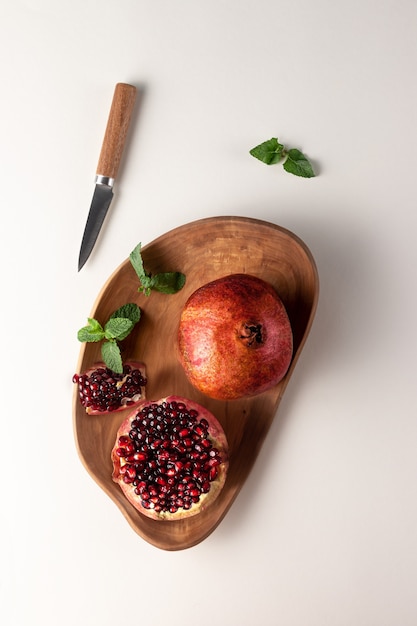 나무 접시 위쪽 전망에 붉은 석류가 잘린 음식 구성. 자연 그림자가 있는 흰색과 회색 배경에 분리된 신선한 민트와 칼