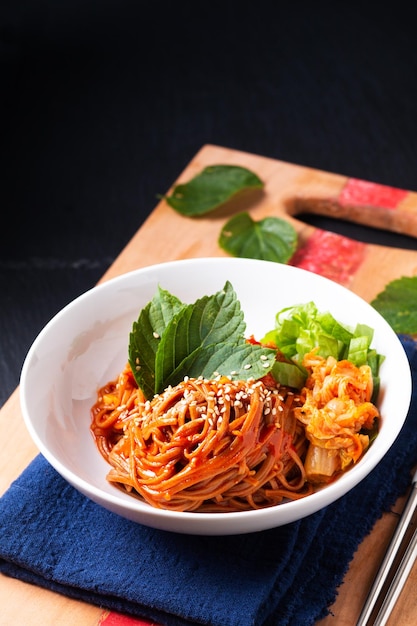 Concetto di cibo coreano piccante freddo noodle di grano saraceno kimchi bibim guksu su tavola di legno e sfondo nero con spazio di copia