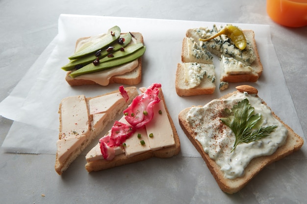 Food concept. Snack concept. Different kinds of colorful sandwiches with rare ingredients and glass of orange juice for having snack or break over white cooking paper.