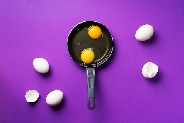 Food concept with two eggs, shells, pan on violet background