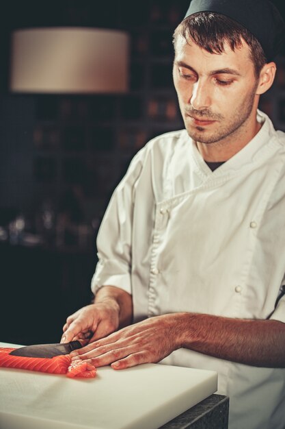 음식 개념 젊은 셰프는 흰색 유니폼을 입은 그가 일하는 레스토랑의 식탁에 연어 생선을 자른다