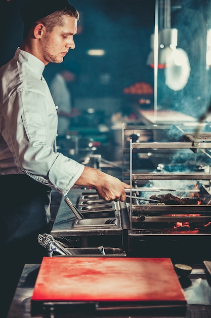 Concetto di cibo. il giovane chef bello in uniforme bianca controlla il grado di tostatura e trasforma la carne