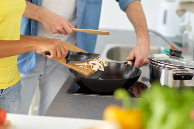 Food, cooking, people, international and healthy eating concept - multiethnic couple with pan frying champignons at home kitchen