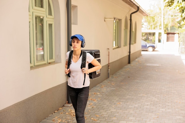 Food courier in headphones walking in street, standing outdoors, front view. Delivery by foot, woman going with customer order in thermal backpack, listening to music, delivering eatery meal