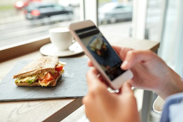 Фото Еда, кулинария, технологии и концепция людей - женские руки со смартфоном фотографируют сэндвич панини в ресторане