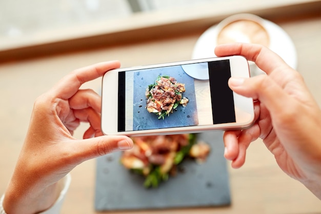 Foto concetto di cibo, cucina, tecnologia e persone - mani di donna con smartphone che fotografano insalata di prosciutto su lastra di pietra al ristorante