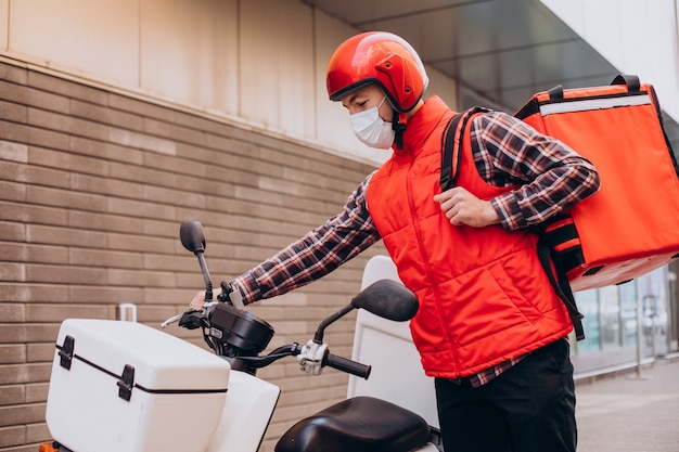 Ragazzo delle consegne di cibo alla guida di uno scooter con scatola con cibo e maschera da indossare