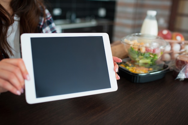 Concetto di consegna del cibo. una giovane donna ordina cibo usando un laptop a casa. sul tavolo ci sono latte, insalate in scatole, carne, cibo, frutta, uova, pane.