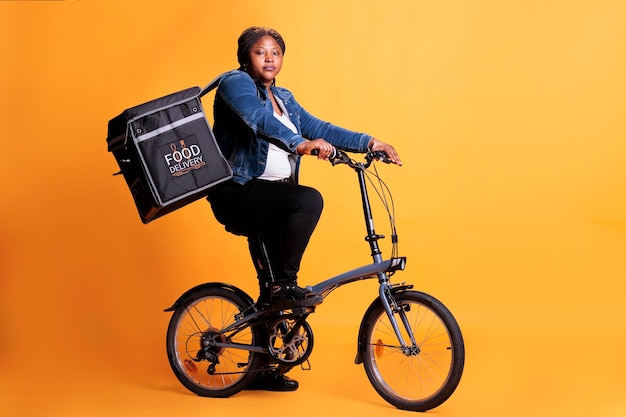 Photo food delivery employee riding bike while carrying takeaway thermal backpack ready to deliver order to client during lunch time, standing over yellow background. food service and transportation