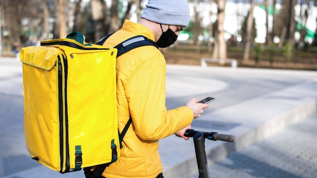 Фото Доставщик еды на скутере в парке с помощью своего смартфона. черная медицинская маска, желтый рюкзак и куртка. зима